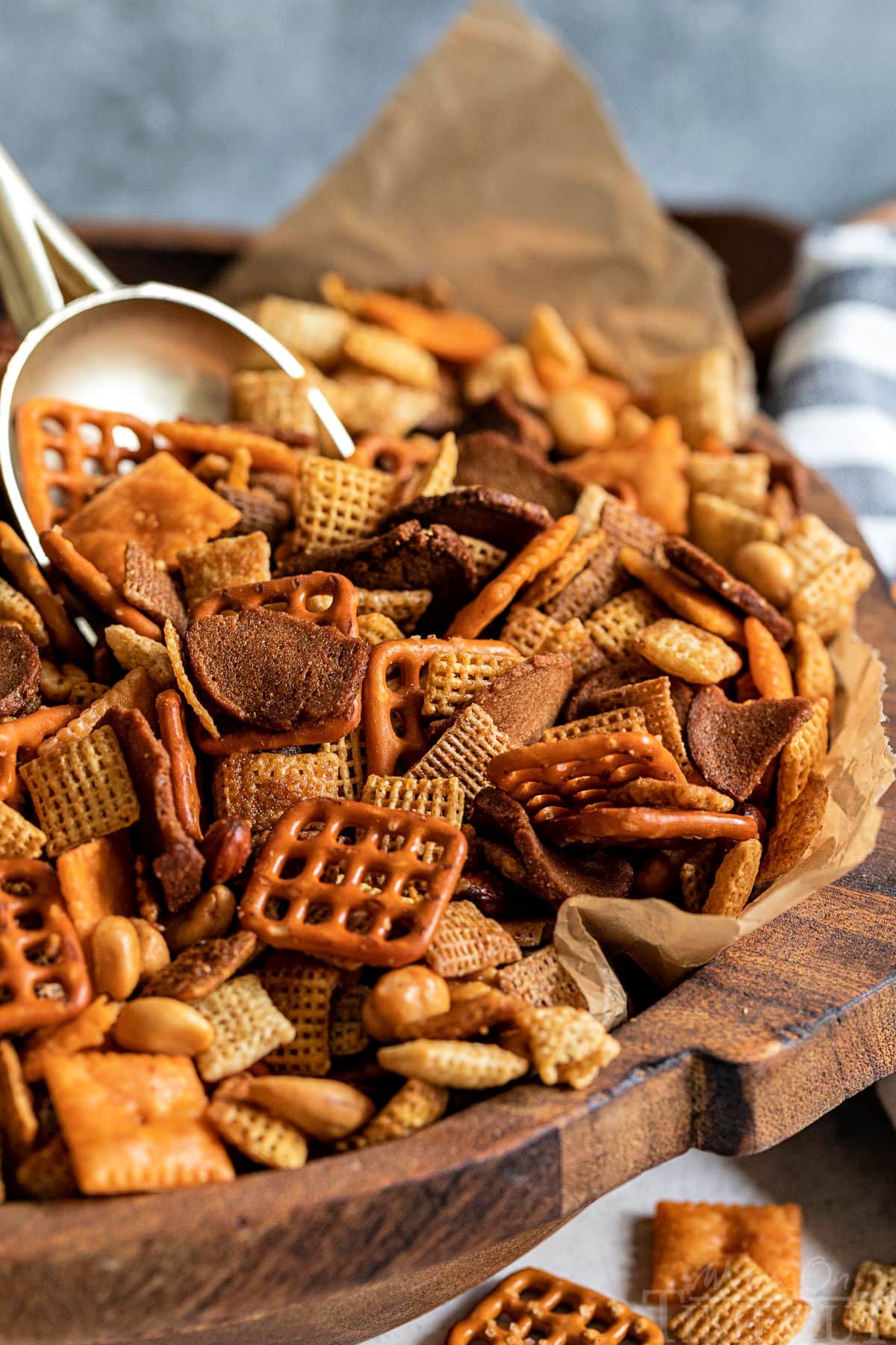 large wood bowl lined with brown parchment paper and filled with chex mix. large gold scoop sticking out of the chex mix recipe.