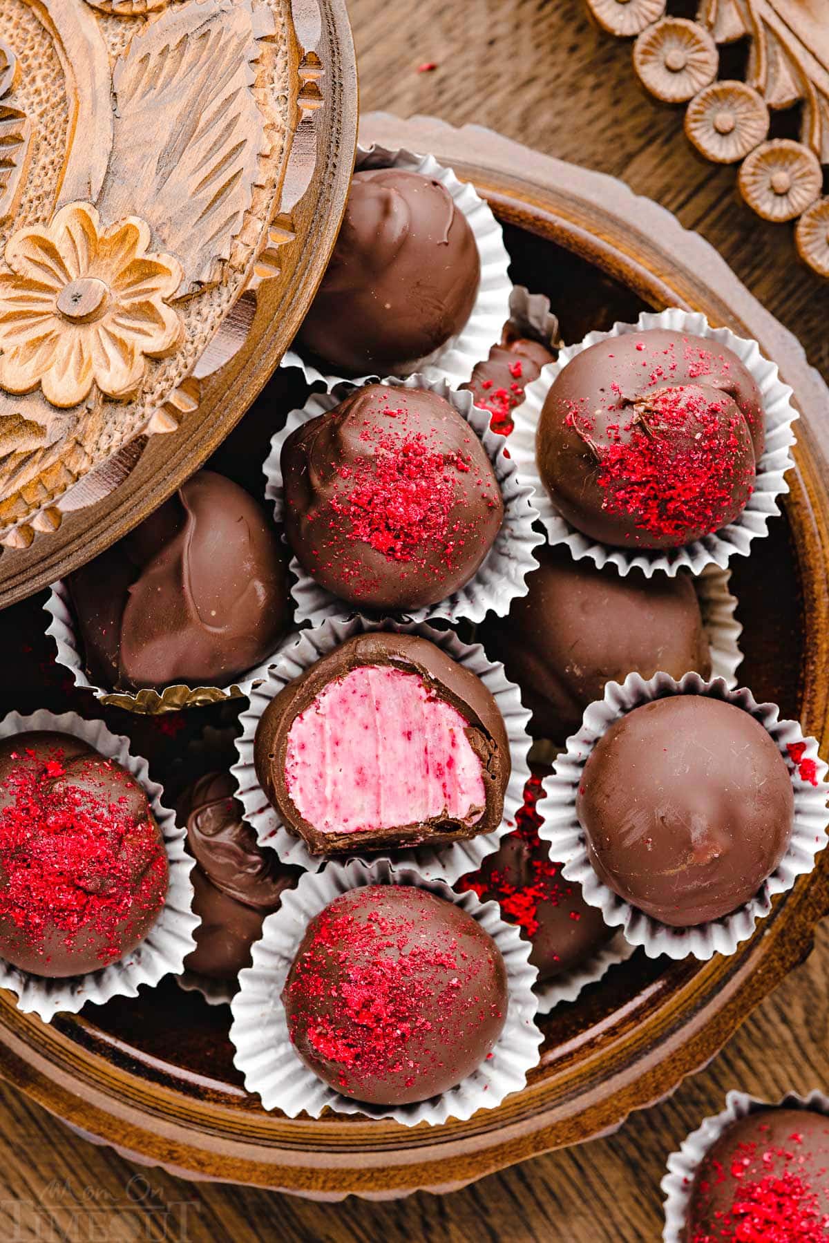 round wooden box filled with raspberry truffles. center truffle has a bite taken out of it. some of the truffles are topped with freeze dried raspberry dust.