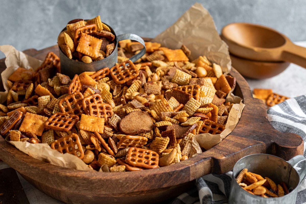 large wooden bowl filled with chex mix with a metal cup in the bowl scooping out the mix.