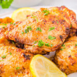 Four chicken thighs cooked in air fryer piled on a white plate and garnished with fresh parsley and lemon slices.