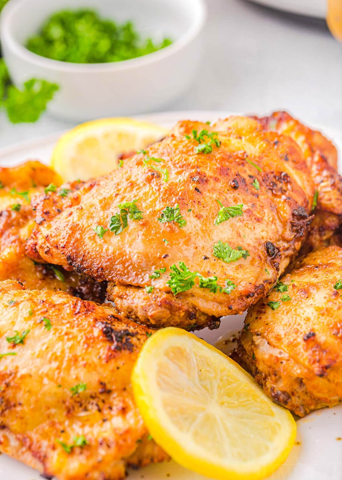 Four chicken thighs cooked in air fryer piled on a white plate and garnished with fresh parsley and lemon slices.