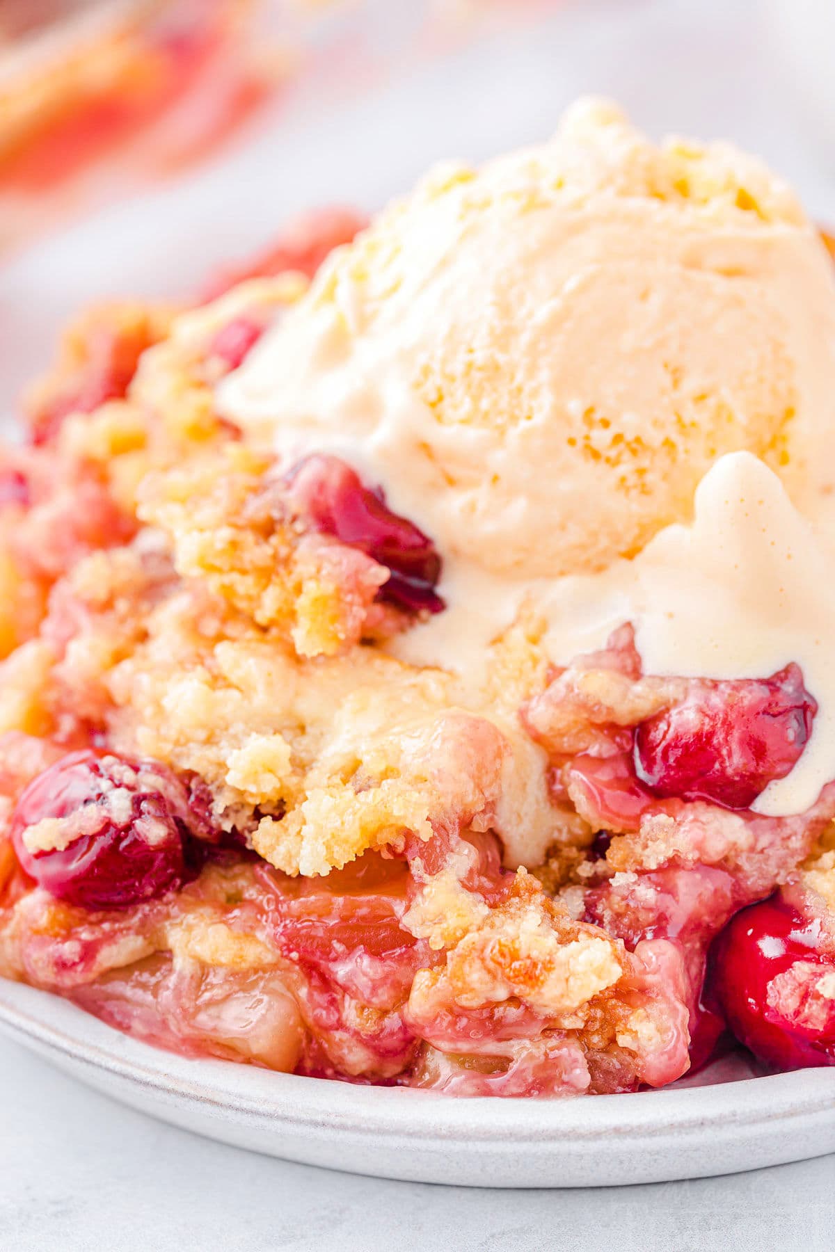 plate filled with cherry pineapple dump cake and topped with a scoop of vanilla ice cream that is just beginning to melt. cherries and pineapple chunks are visible in the cake.