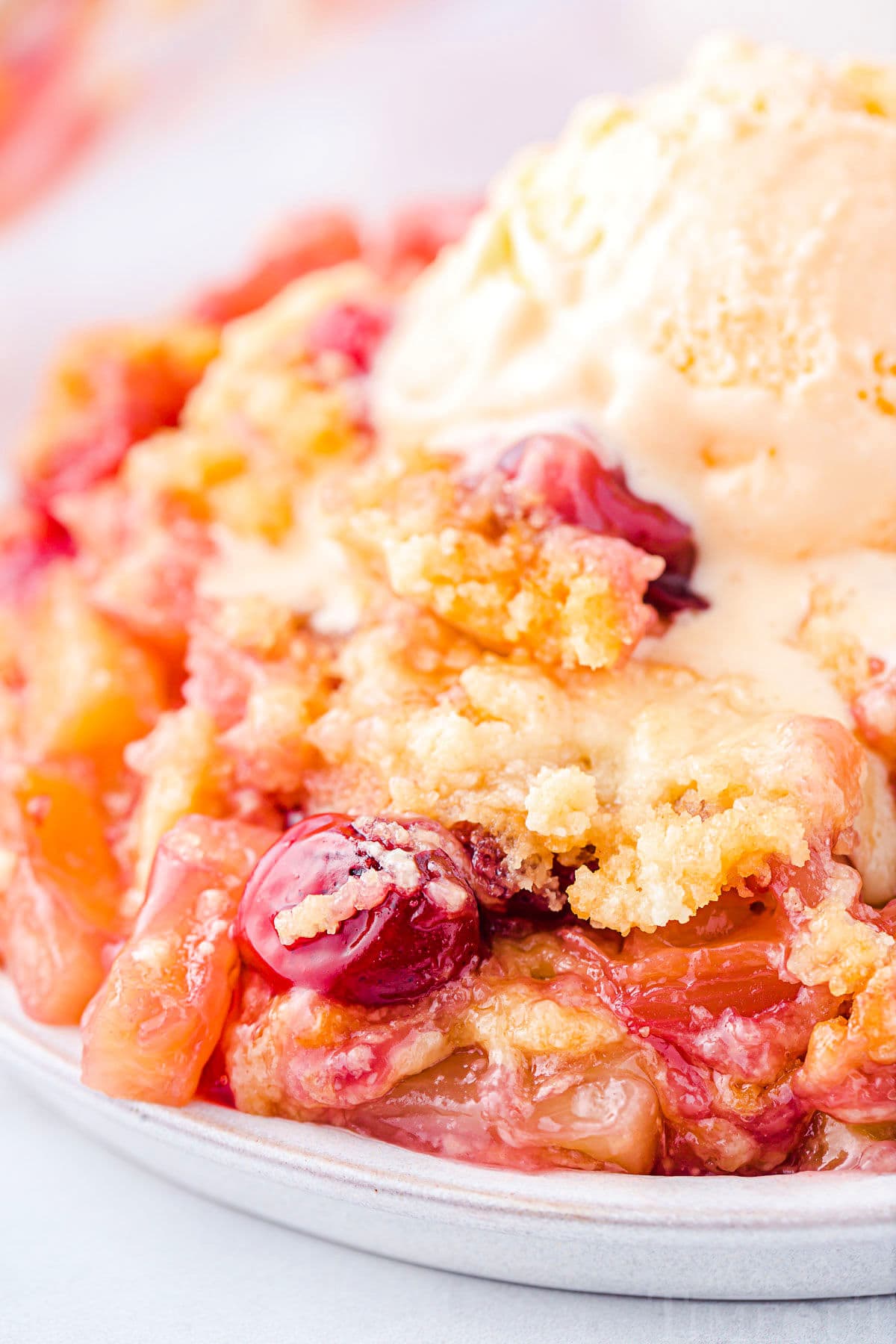 plate filled with cherry pineapple dump cake and topped with a scoop of vanilla ice cream that is just beginning to melt. cherries and pineapple chunks are visible in the cake.