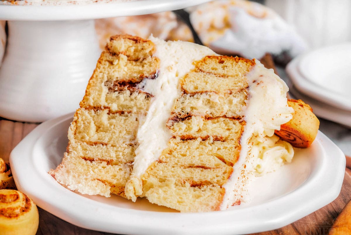 large slice of cinnamon roll layer cake on a white plate with a white cake stand in the background.