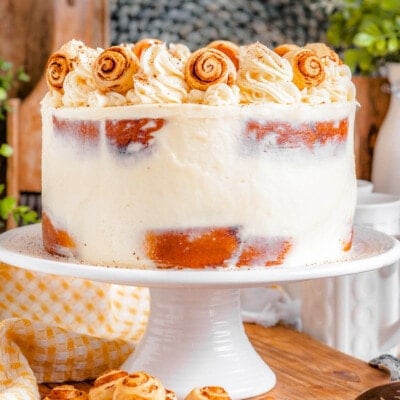 cake made with cinnamon rolls on a white cake stand and topped with cream cheese frosting and mini cinnamon rolls.