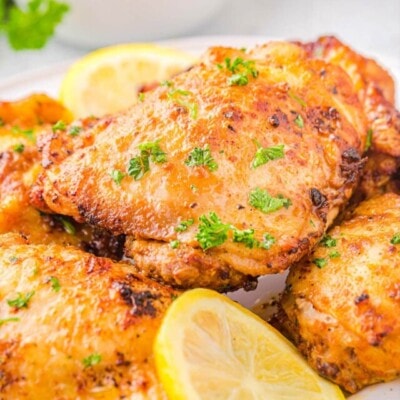 Four chicken thighs cooked in air fryer piled on a white plate and garnished with fresh parsley and lemon slices.