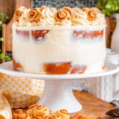 cake made with cinnamon rolls on a white cake stand and topped with cream cheese frosting and mini cinnamon rolls.