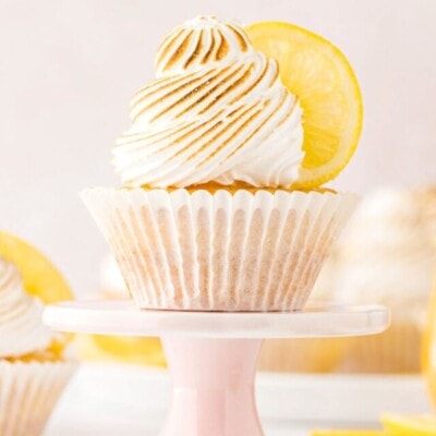 two cupcakes topped with meringue and filled with lemon curd. one cupcake is sitting on a pastel pink cupcake stand. jar of lemon curd in background.