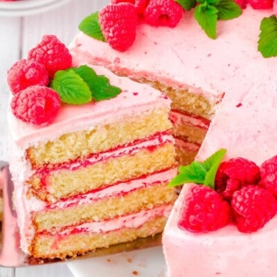 slice being removed from a vanilla cake with raspberry buttercream. cake is decorated with fresh raspberry and mint leaves.