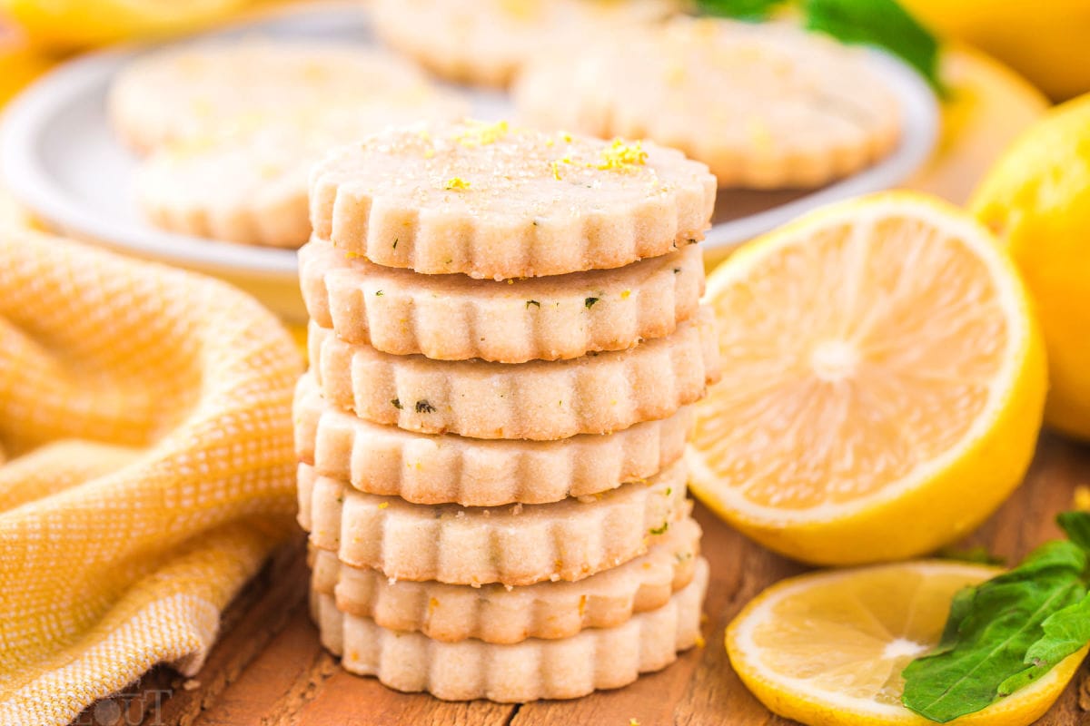 stack of lemon basil shortbread cookies stacked in front of a plate of cookies with lemons cut in half and yellow napkin around the plate.