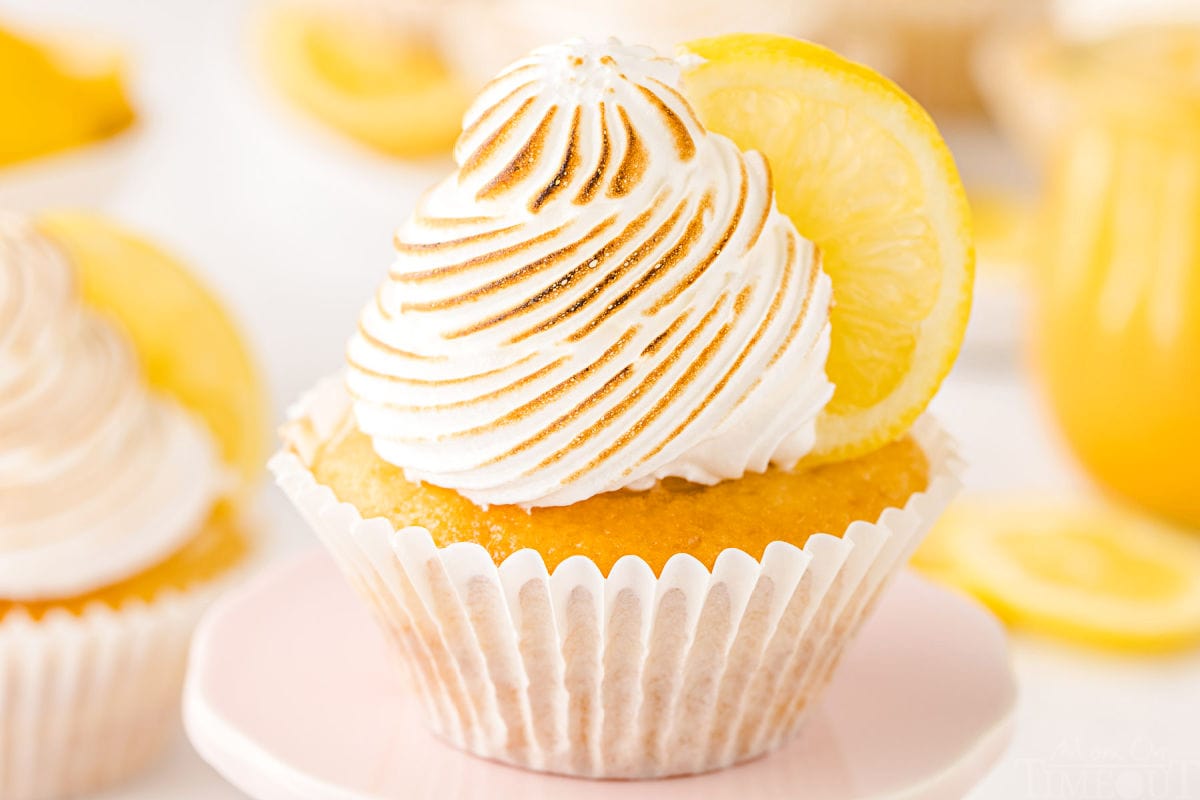 cupcake topped with meringue and filled with lemon curd. one cupcake is sitting on a pastel pink cupcake stand. jar of lemon curd in background.