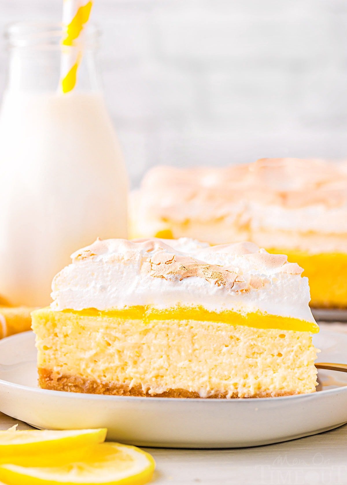 one slice of lemon meringue cheesecake sitting on a white plate with the rest of the cheesecake seen in the background next to a small bottle of milk. lemon slices near the front of the plate.