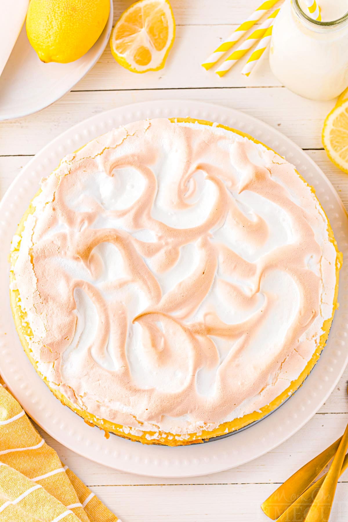 top down look at lemon cheesecake with a meringue topping. the meringue is beautifully swirled with golden brown tips. Lemon slices and wedges are around the cheesecake.