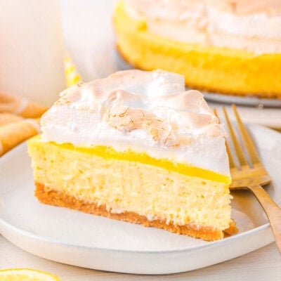 one slice of lemon meringue cheesecake sitting on a white plate with the rest of the cheesecake seen in the background next to a small bottle of milk. lemon slices near the front of the plate.