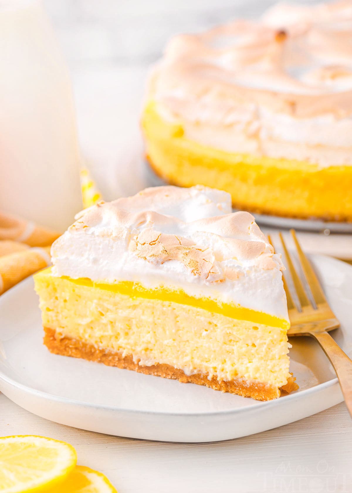 one slice of lemon meringue cheesecake sitting on a white plate with the rest of the cheesecake seen in the background next to a small bottle of milk. lemon slices near the front of the plate.