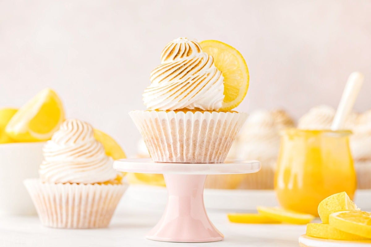 two cupcakes topped with meringue and filled with lemon curd. one cupcake is sitting on a pastel pink cupcake stand. jar of lemon curd in background.