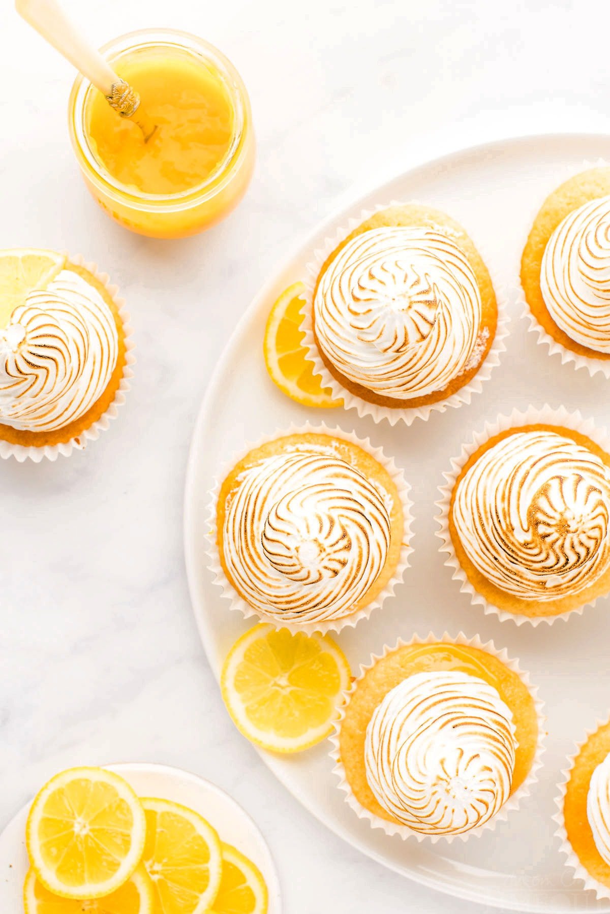white plate with six lemon meringue cupcakes on it as well as a bowl of lemons cut into wedges.