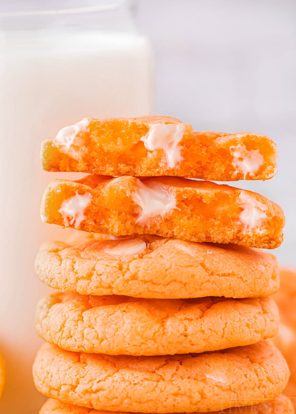 five orange cookies stacked with a glass of milk in background. top cookie is broken in half and you can see the melty white chocolate chips in the cookies.