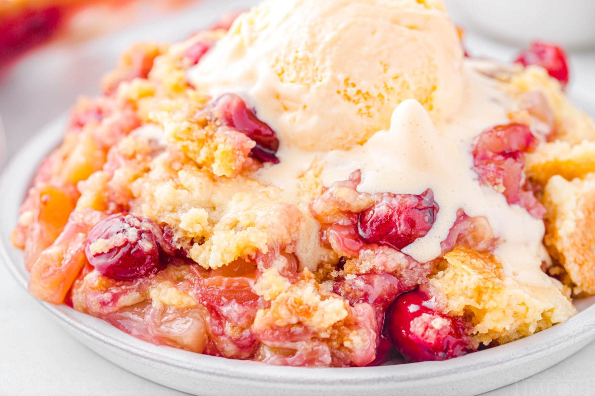 shallow plate filled to the brim with a generous serving of pineapple cherry dump cake. the cake is topped with a scoop of vanilla ice cream that is melting and dripping into the cake.