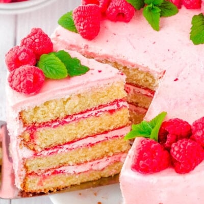 slice being removed from a vanilla cake with raspberry buttercream. cake is decorated with fresh raspberry and mint leaves.
