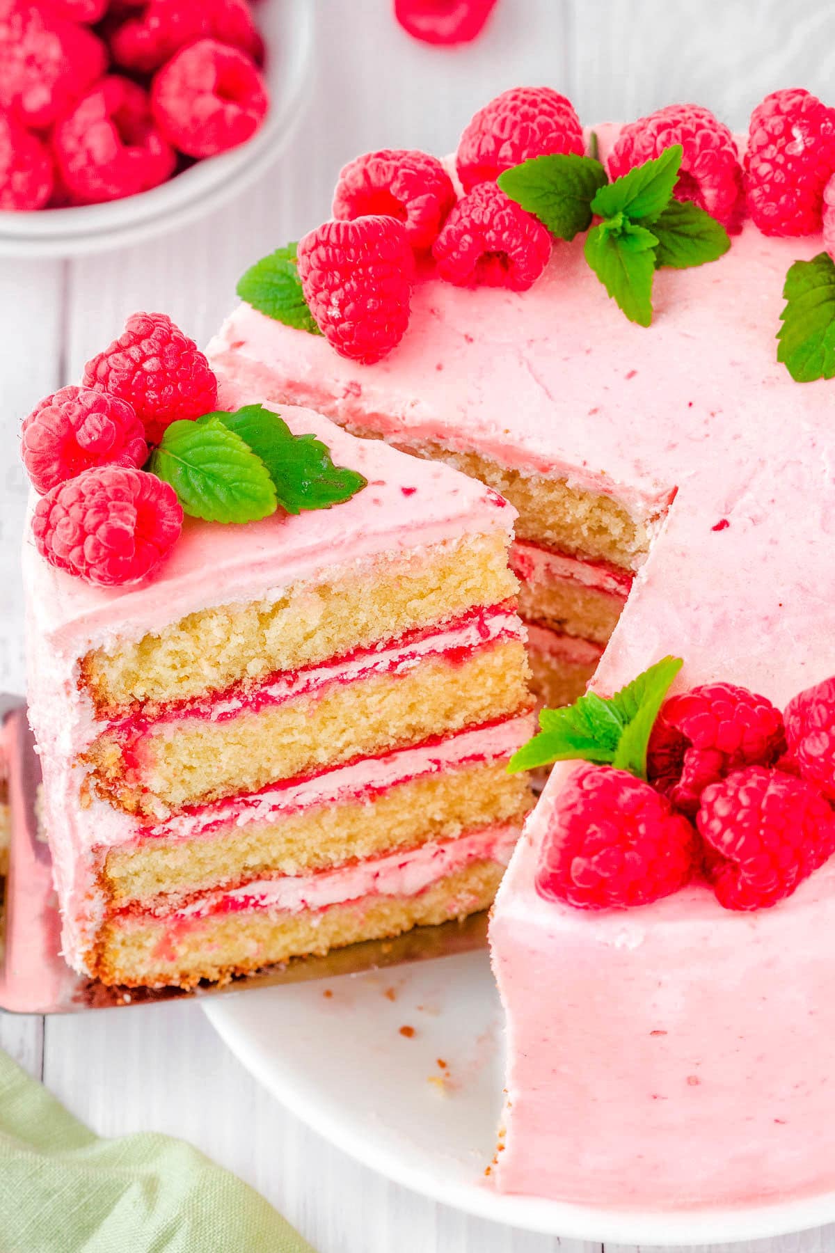 slice being removed from a vanilla cake with raspberry buttercream. cake is decorated with fresh raspberry and mint leaves.