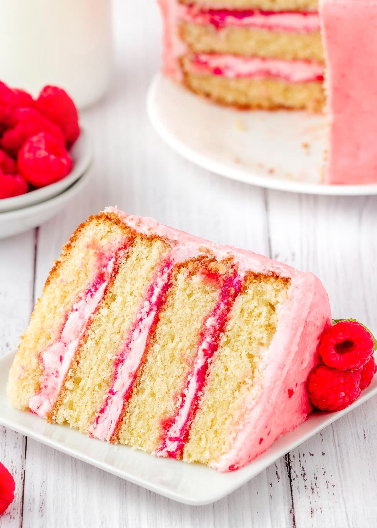 slice of vanilla raspberry cake on a white square plate in front of the rest of the cake.