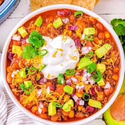 top down look at a generous serving of taco soup in a white bowl topped with avocado, sour cream and cheese. tortilla chips and other toppings can be seen around the bowl.