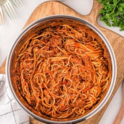 spaghetti in instant pot ready to be served sitting on a wood cutting board with parsley and parmesan next to the board.