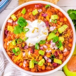 top down look at a generous serving of taco soup in a white bowl topped with avocado, sour cream and cheese. tortilla chips and other toppings can be seen around the bowl.