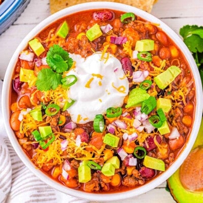 top down look at a generous serving of taco soup in a white bowl topped with avocado, sour cream and cheese. tortilla chips and other toppings can be seen around the bowl.