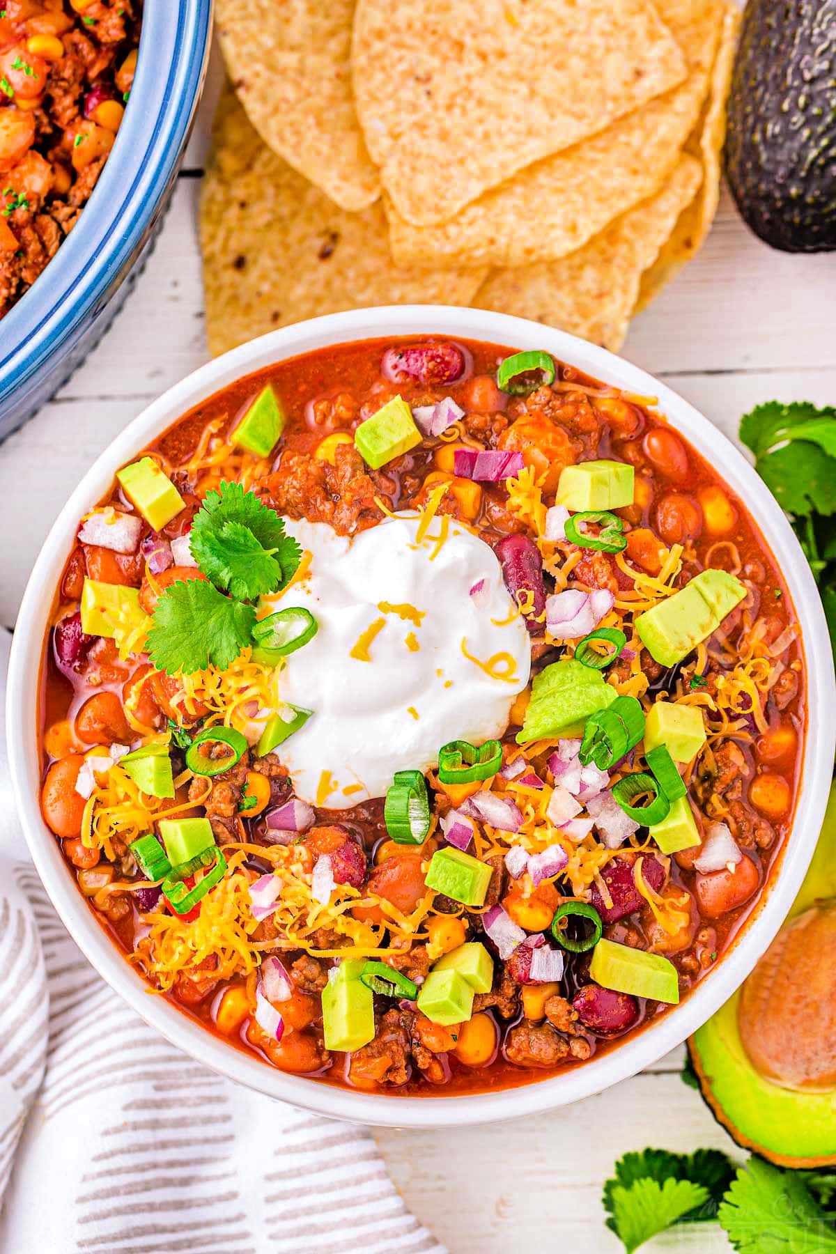 top down look at a generous serving of taco soup in a white bowl topped with avocado, sour cream and cheese. tortilla chips and other toppings can be seen around the bowl.