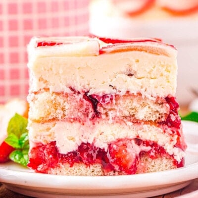 front look at layered strawberry tiramisu serving on a white plate. baking dish and pink checked bowl can be seen in the background.