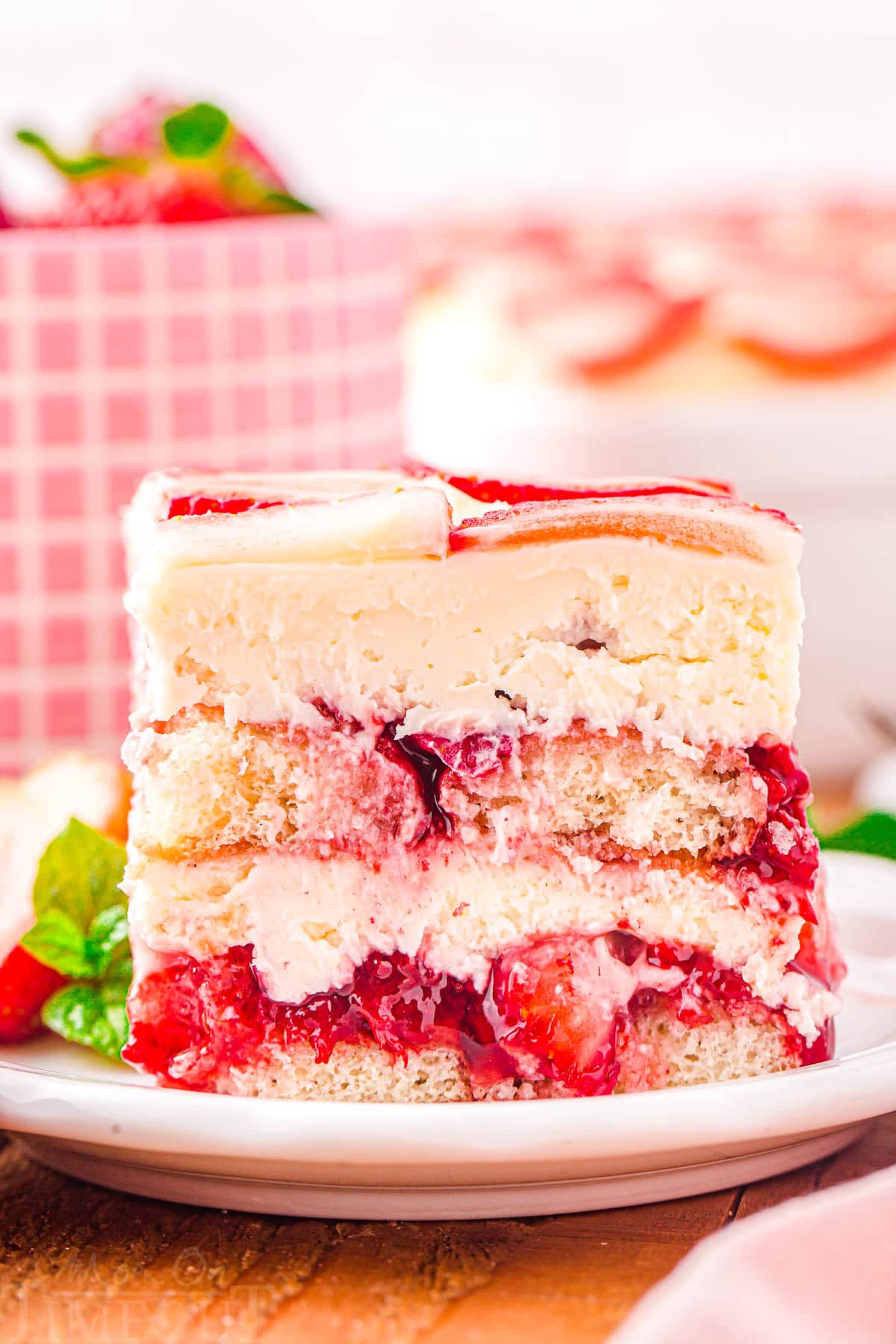 front look at layered strawberry tiramisu serving on a white plate. baking dish and pink checked bowl can be seen in the background.