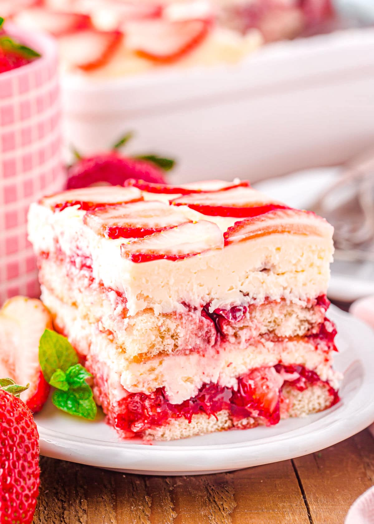 piece of strawberry tiramisu in a white plate with fresh strawberries on top and around the plate.