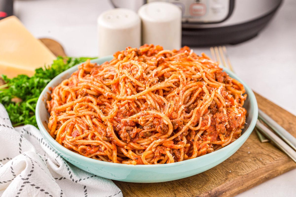 aqua colored serving bowl filled with spaghetti sitting on a wood board next to a white towel in front of an instant pot.