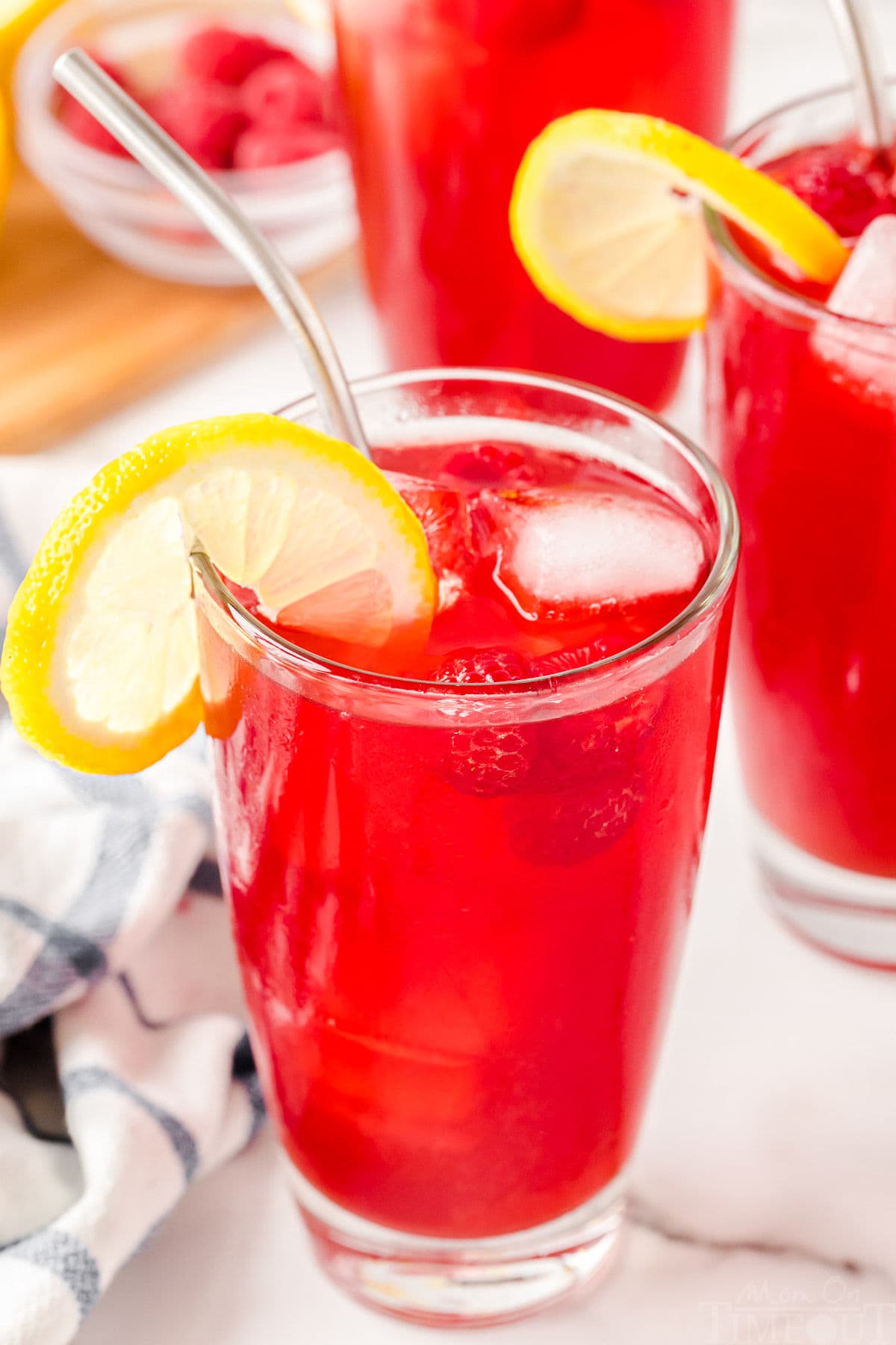 top down angled view of a glass filed with vibrantly colored raspberry iced tea topped with a lemon slice. a stainless steel straw is inserted into the glass and two more filled glasses can be seen in the background.
