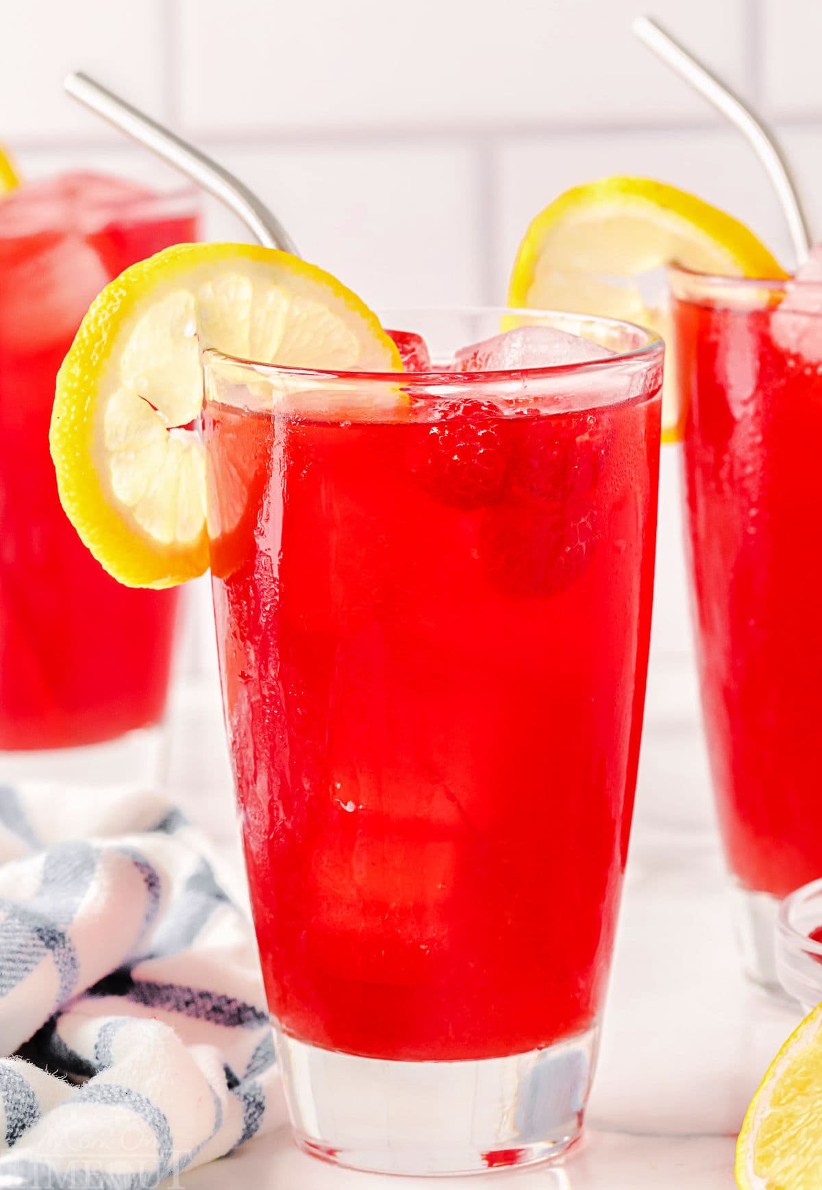 large tall glass filled with raspberry iced tea and topped with a lemon slice. two more glasses can be seen in the background.