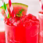 close up view of a tall clear glass filled to the top with watermelon slushie. Topped with a sprig of mint and watermelon wedge with two paper straws inserted.