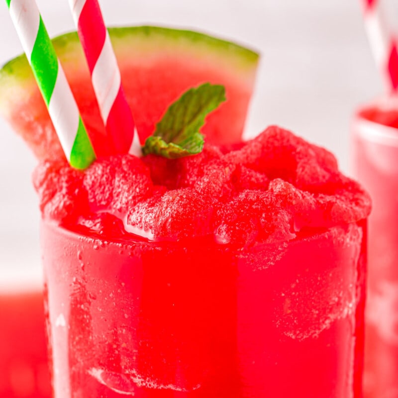 close up view of a tall clear glass filled to the top with watermelon slushie. Topped with a sprig of mint and watermelon wedge with two paper straws inserted.