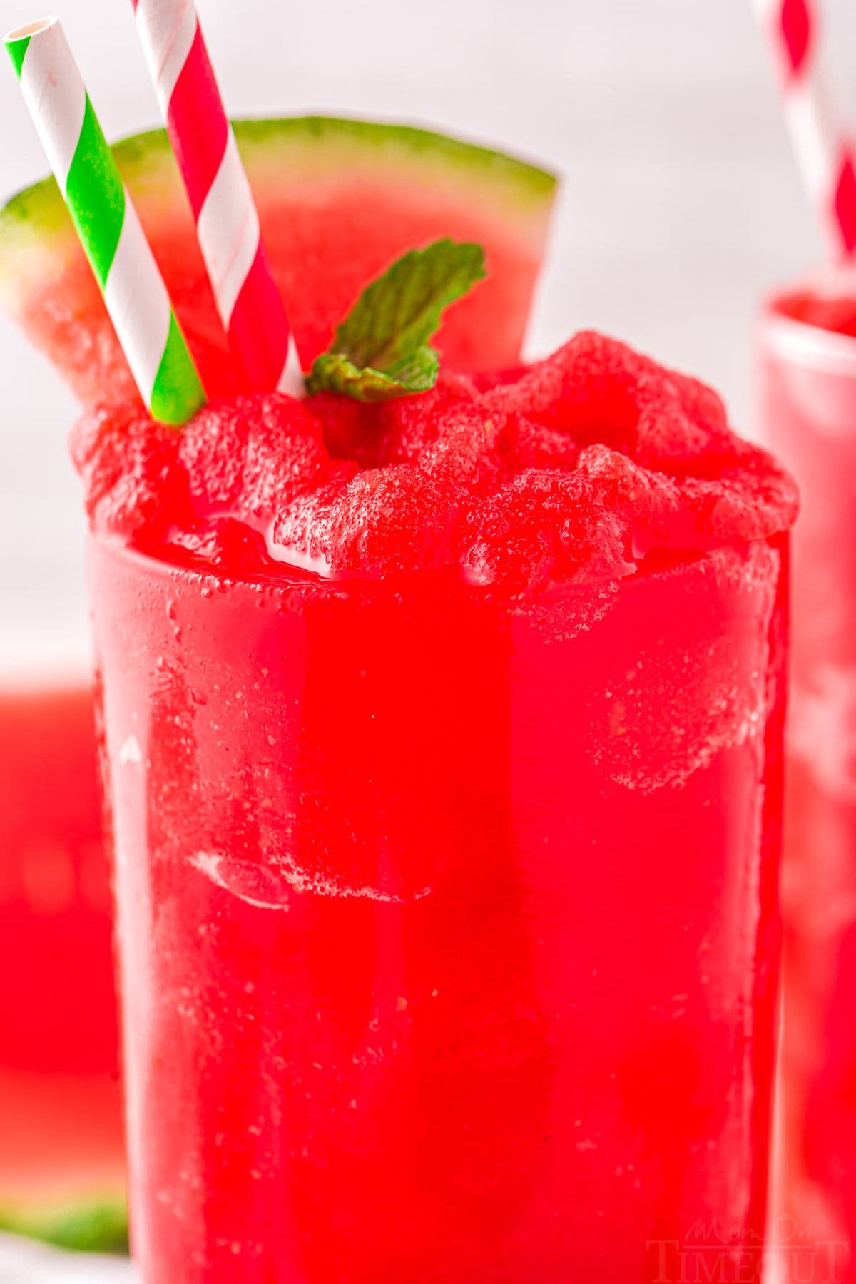 close up view of a tall clear glass filled to the top with watermelon slushie. Topped with a sprig of mint and watermelon wedge with two paper straws inserted.
