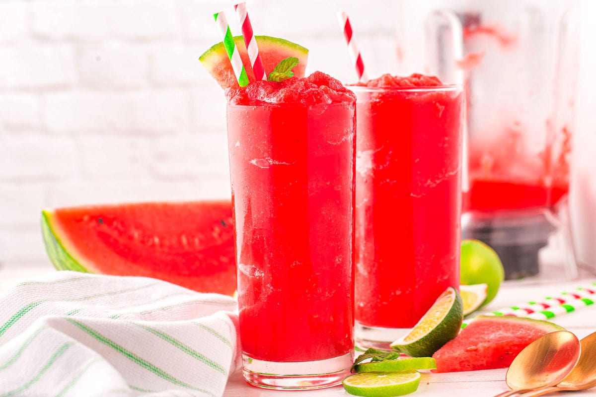 two tall glasses filled with bright red watermelon slushie and topped with a watermelon wedge. Wedge of watermelon can be seen in the background along with the blender.
