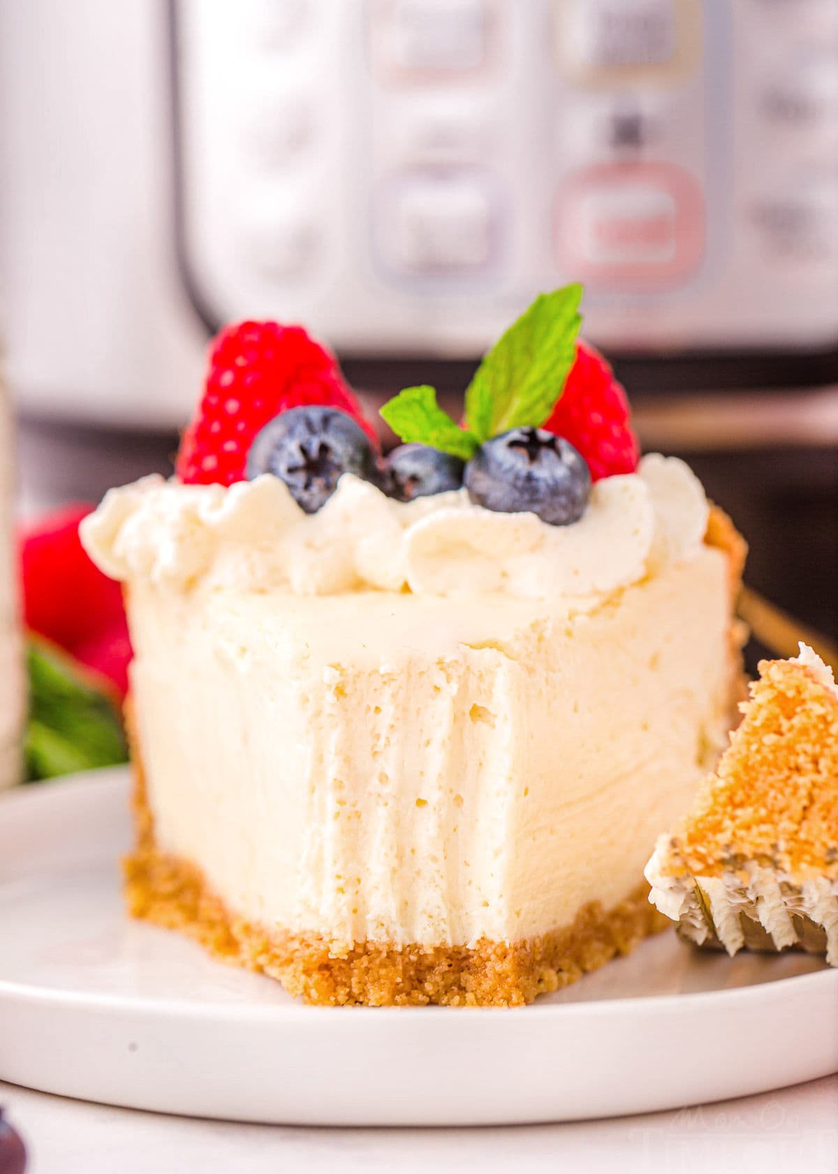 piece of instant pot cheesecake on a small round white plate with a forkful removed. cheesecake is topped with fresh berries and whipped cream.