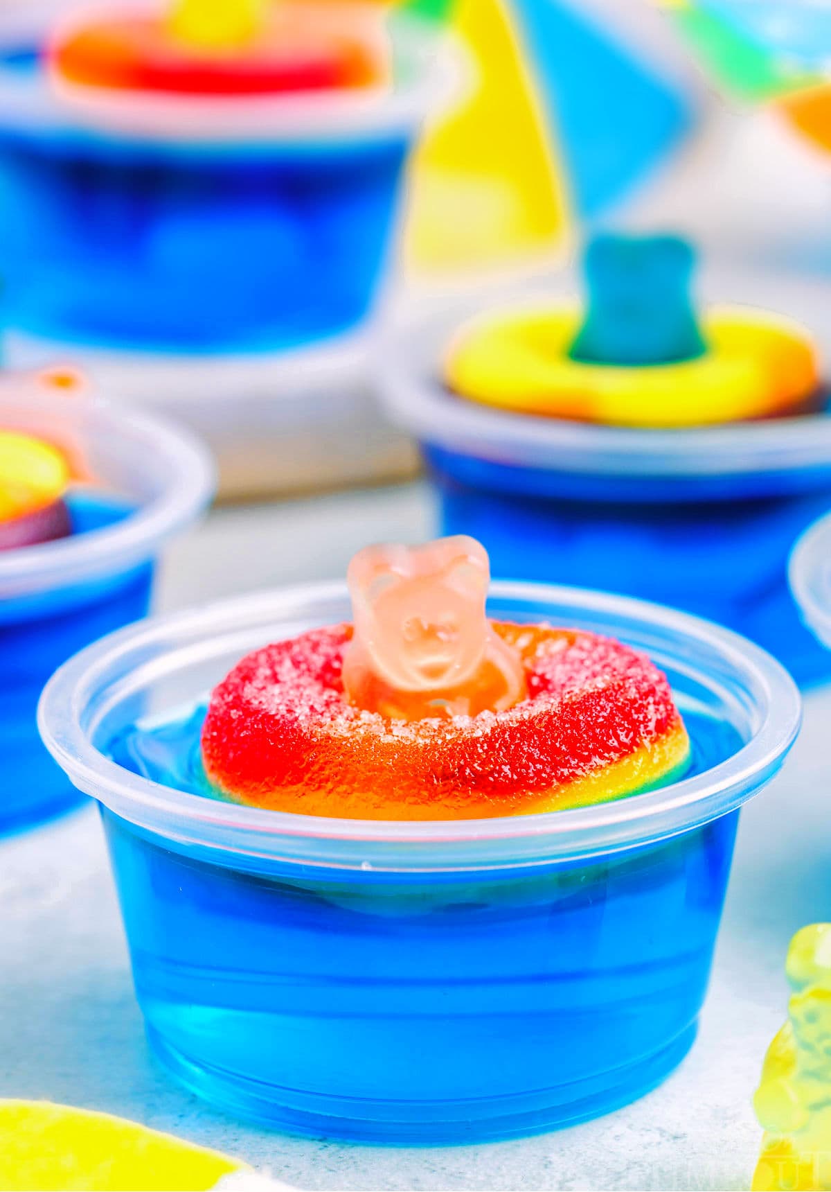 blue pool party jello shot with gummi bear sitting on white surface. more jello shots can be seen in the background.