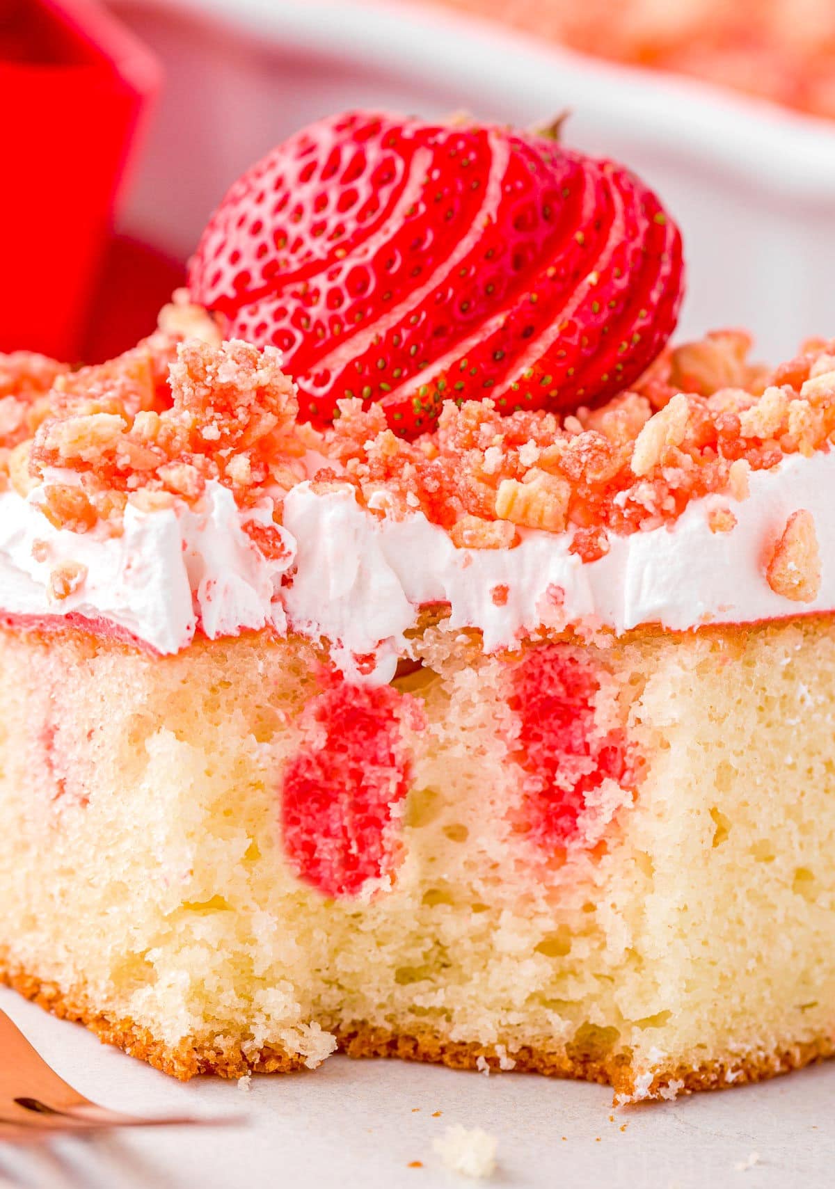 piece of strawberry crunch cake on plate with one forkful removed. close up shows the red jello that has been added to the poke cake.