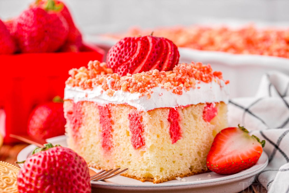 serving of strawberry poke cake on small white round plate topped with fresh sliced strawberry and crunch topping. rest of cake can be seen in background.