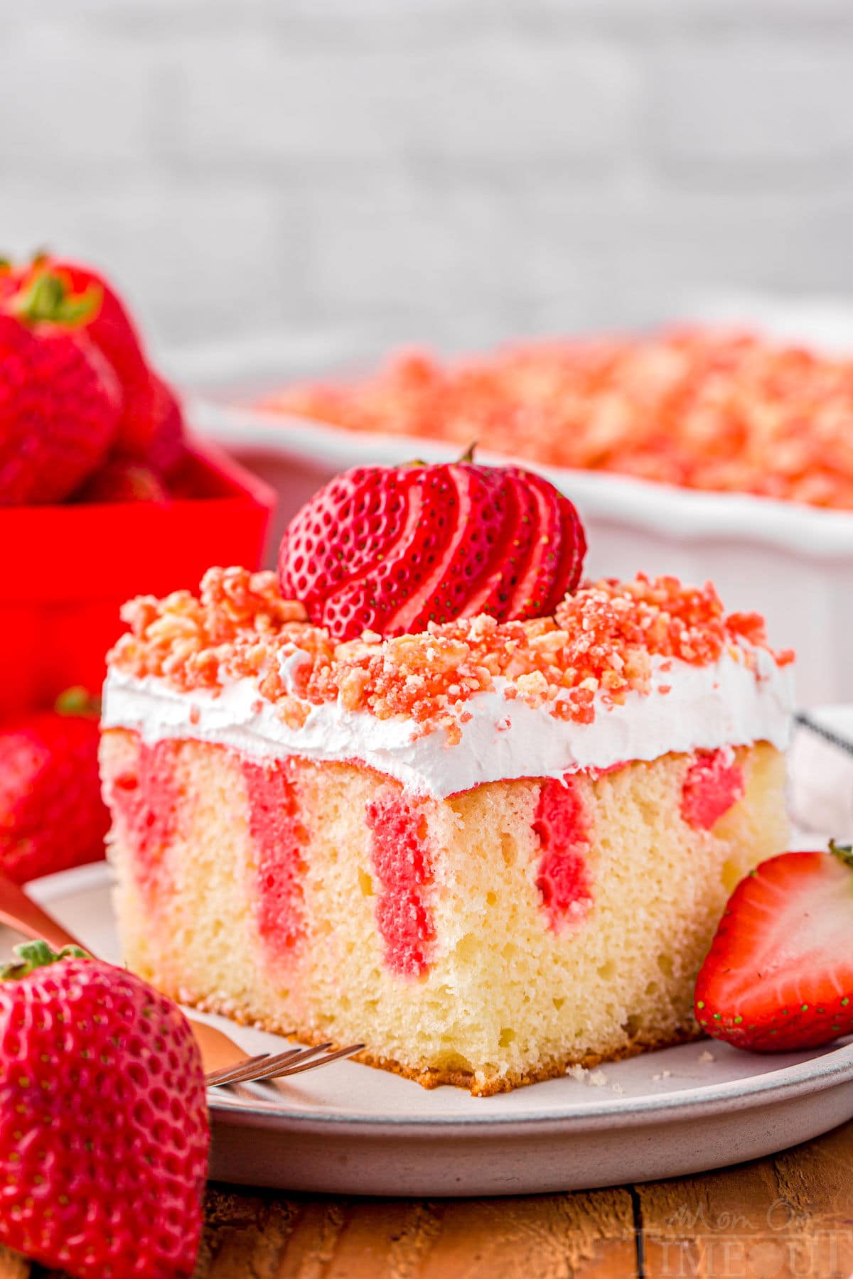 serving of strawberry poke cake on small white round plate topped with fresh sliced strawberry and crunch topping. rest of cake can be seen in background.