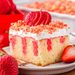serving of strawberry poke cake on small white round plate topped with fresh sliced strawberry and crunch topping. rest of cake can be seen in background.