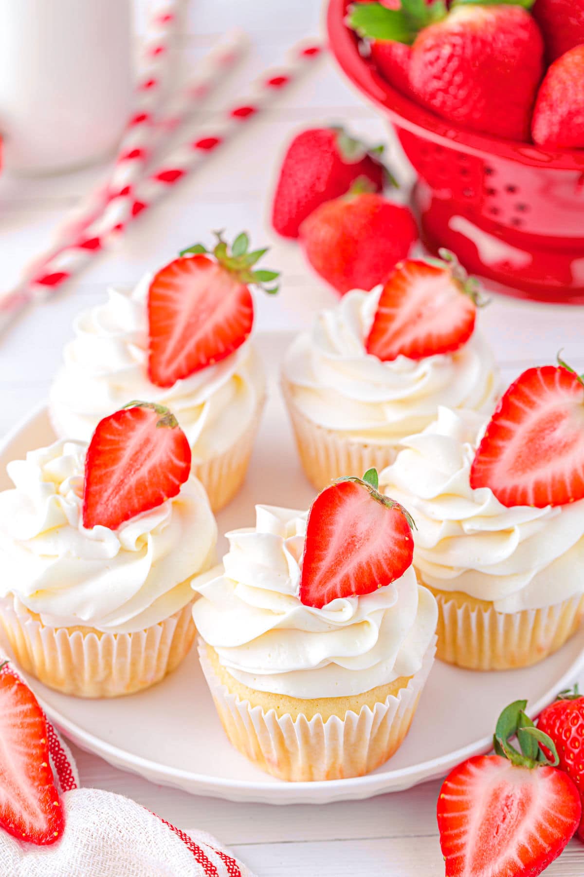 top down view of five vanilla cupcakes topped with vanilla buttercream and filled with strawberry jam on white plate. Each cupcakes is topped with half a strawberry.