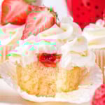 vanilla cupcake with strawberry filling and buttercream frosting. topped with half of a fresh strawberry. cupcake has a bite taken so you can see the strawberry jam on the inside.