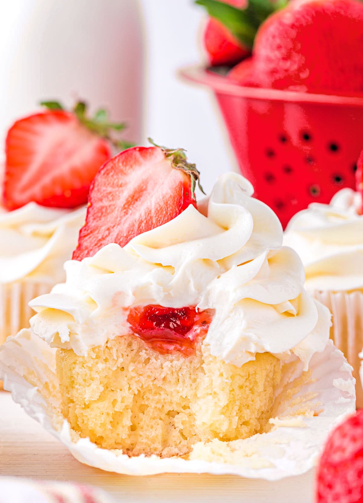 vanilla cupcake with strawberry filling and buttercream frosting. topped with half of a fresh strawberry. cupcake has a bite taken so you can see the strawberry jam on the inside.